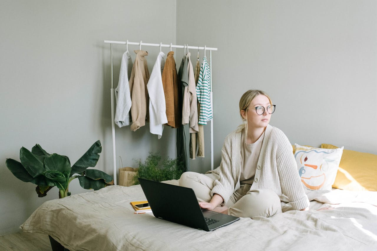 hero-img-02 Adult woman in eyeglasses working from home on a laptop, sitting on a bed with clothes rack nearby.